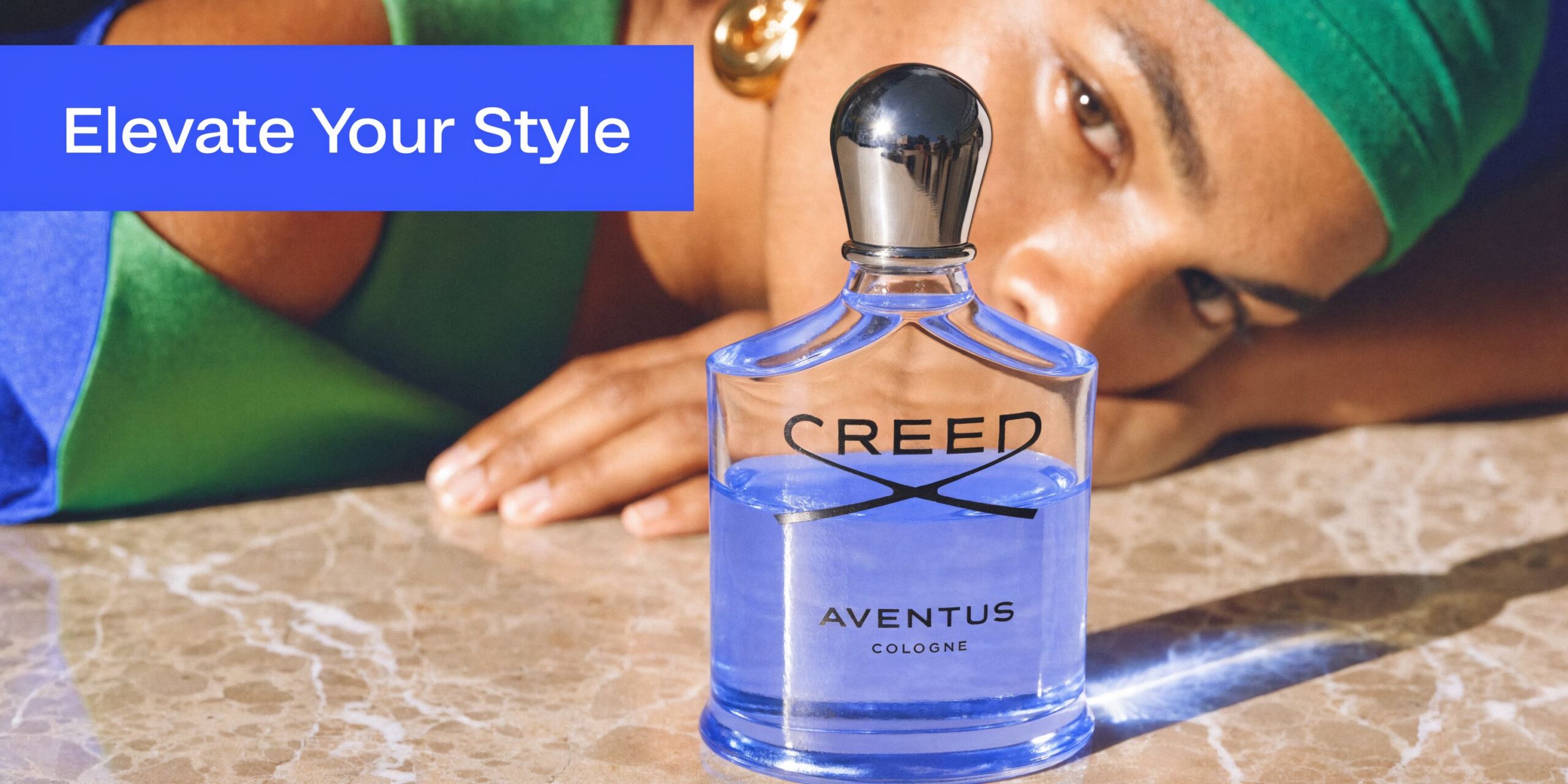 A glass bottle of Creed Aventus Cologne sits on a marble surface near a person lying down.
